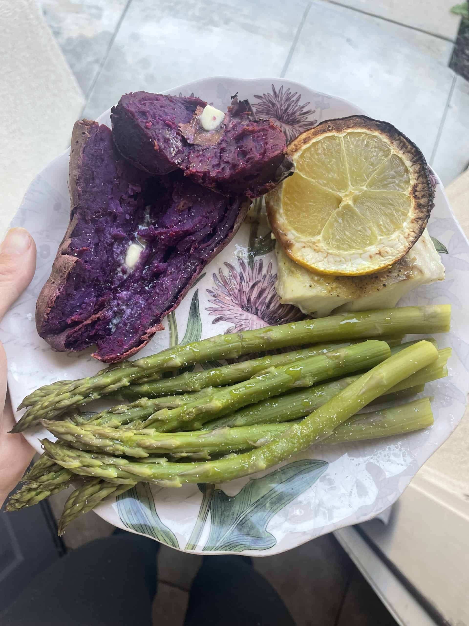 a baked purple sweet potato with asparagus and a lemon slice over a piece of white fish on a decorative plate