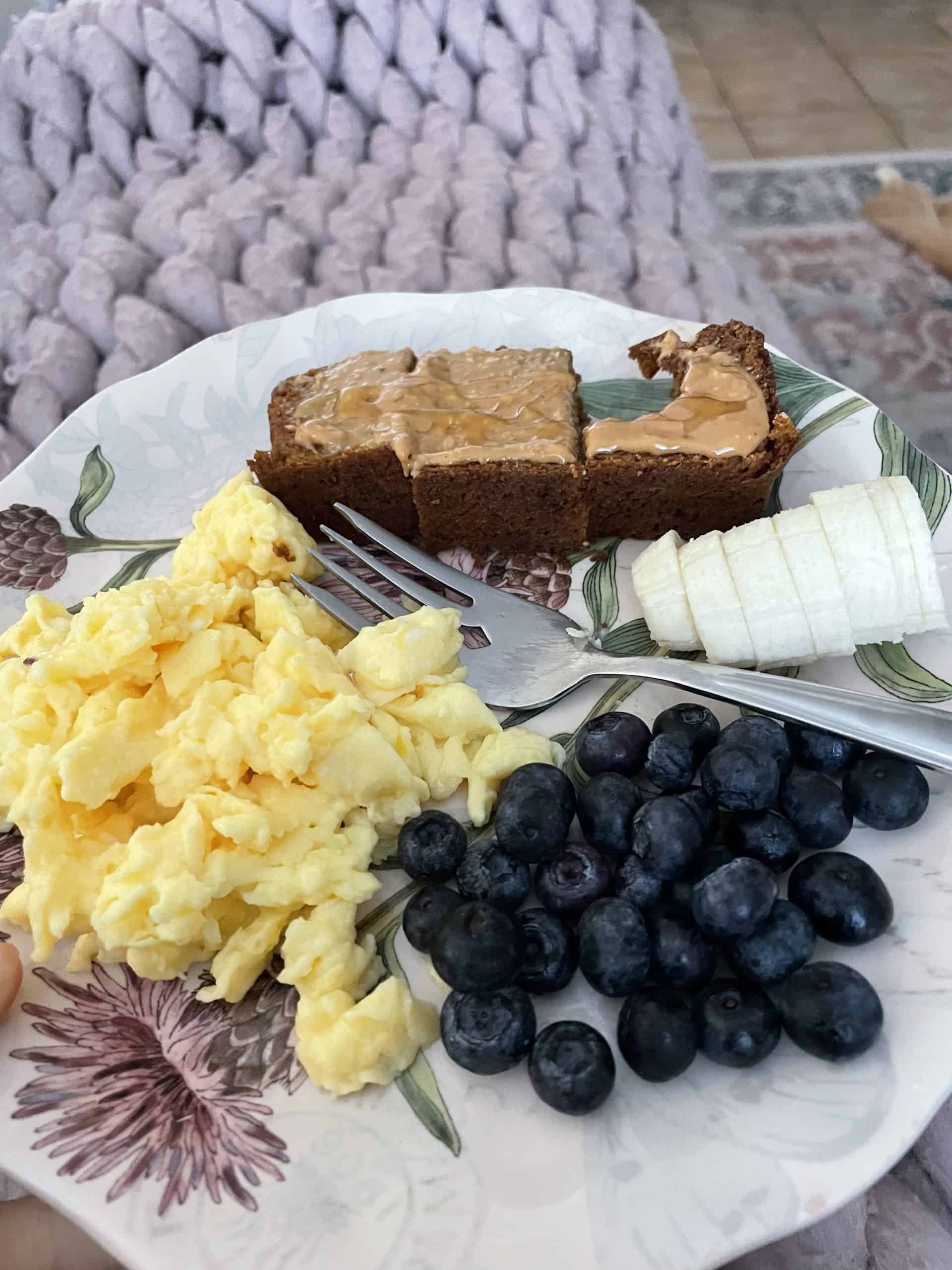 a plate with scrambled eggs, blueberries, banana slices and pumpkin bread on a purple knit blanket