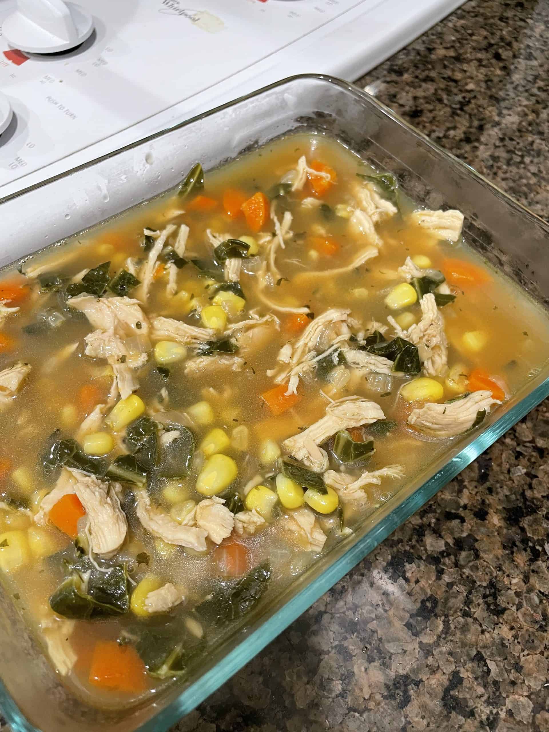 soup in a square glass bowl with shredded chicken, collard greens, corn and diced carrots