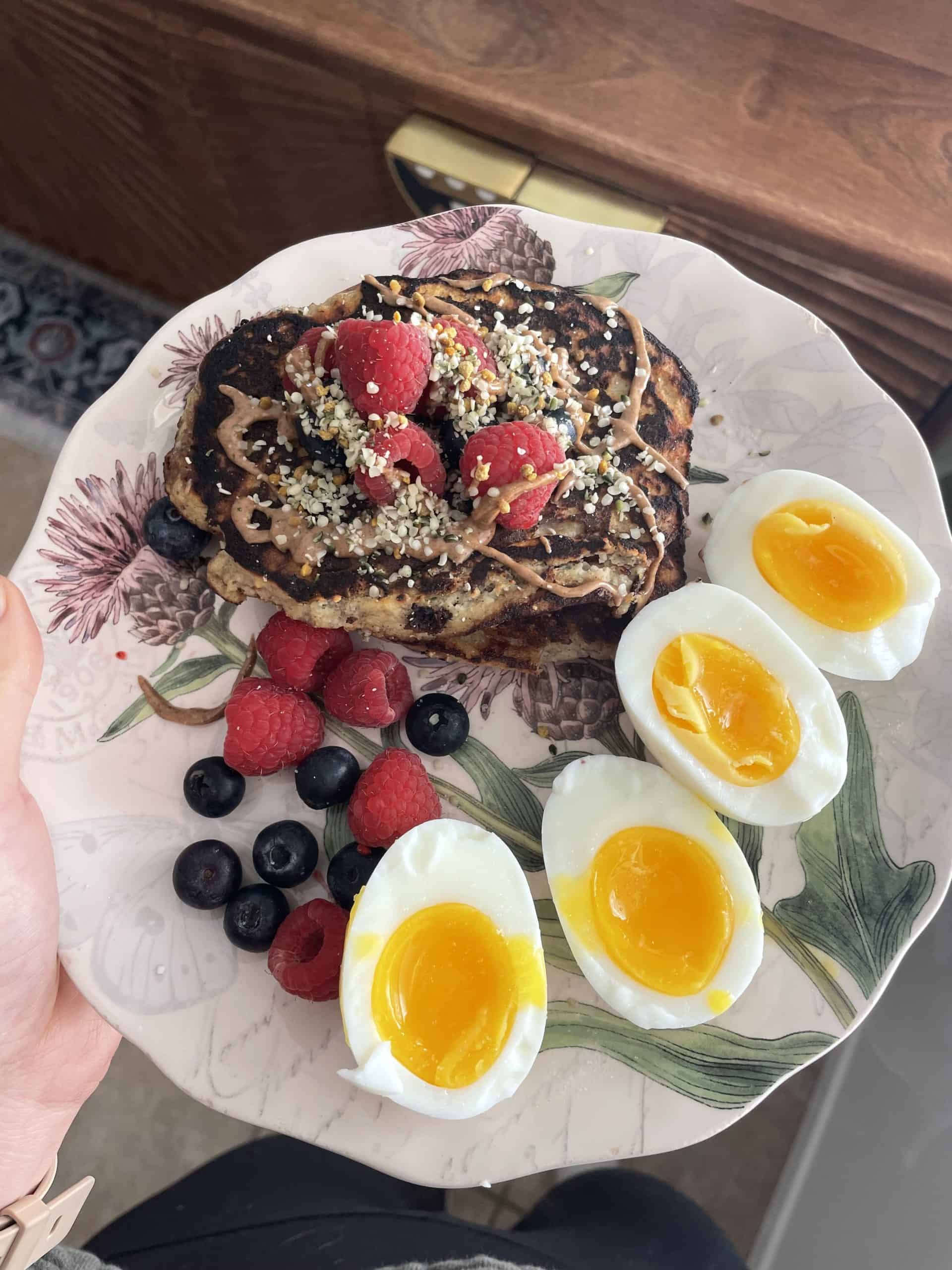 plate of food with pancakes covered with peanut butter, raspberries and hemp seeds and soft boiled eggs on the side